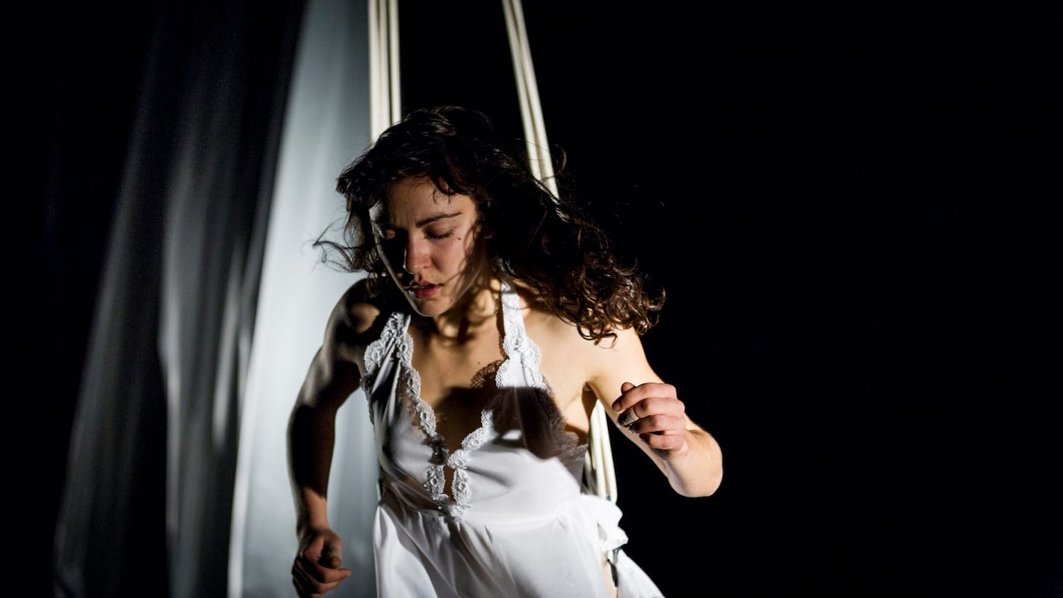 performer with long loose dark hair, wears a short floaty white sleeveless dress in an indoor darkened stage, they are suspended mid-air by ropes