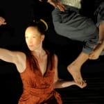 A dancer in a sparkling orange outfit flexes her right arm while holding the foot of another dancer, suspended from above, in her left hand, all set against a dark background.