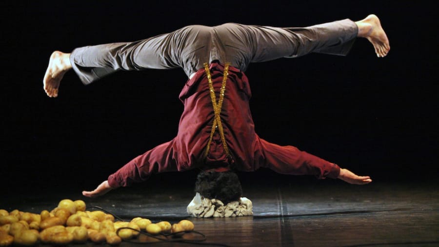 A performer balances upside down on his head, supported by a pile of stones, with potatoes scattered on the ground nearby.