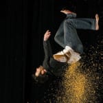 A man in a black jacket and jeans leaps backward, creating a cloud of golden dust against a dark background.