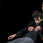 Two individuals in dark clothing strike a dynamic pose against a black background. One stands behind, holding the other securely in both hands.