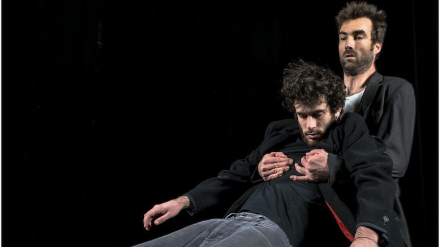 Two individuals in dark clothing strike a dynamic pose against a black background. One stands behind, holding the other securely in both hands.