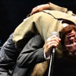 Two performers are on stage - one in a black suit, holding a microphone, and the other in an olive jacket, lying backward on the first's shoulders, obscuring their face."