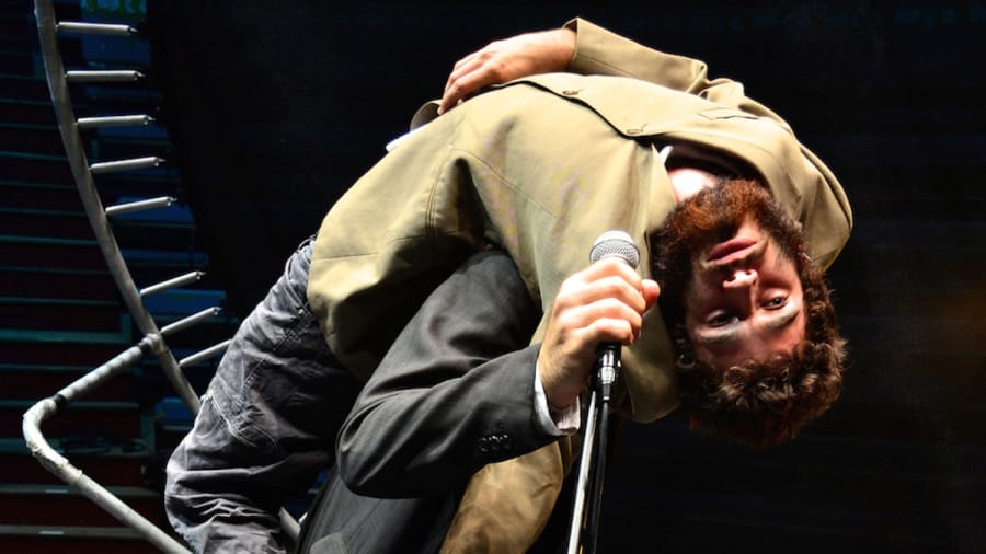 Two performers are on stage - one in a black suit, holding a microphone, and the other in an olive jacket, lying backward on the first's shoulders, obscuring their face."