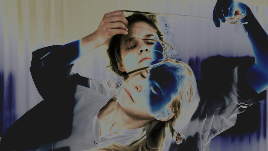 Two performers with closed eyes, one standing behind the other, as the one in the back holds a strip of film in front of their face, creating a surreal effect with inverted colours.