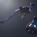 Two acrobats perform a dynamic mid-air stunt in a dimly lit room, showcasing strength, balance, and precision.