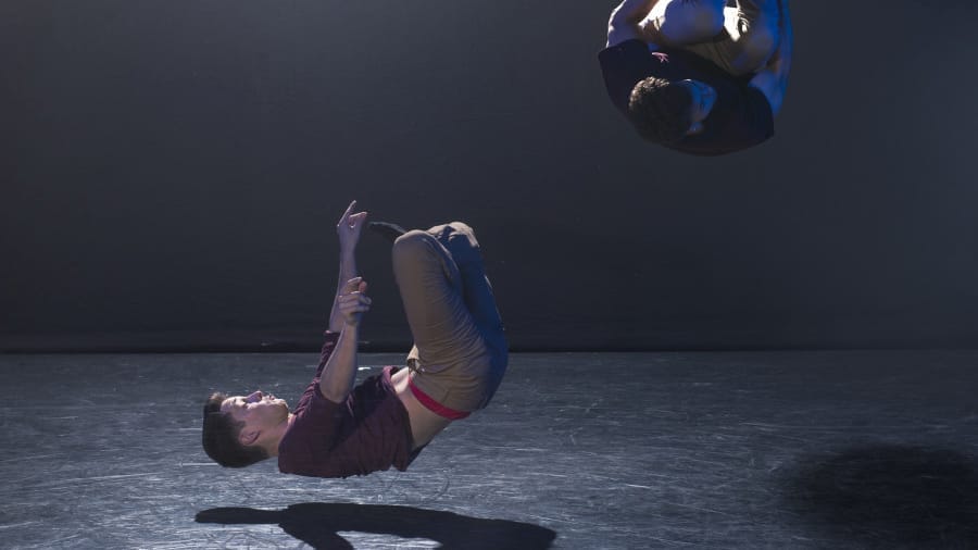 Two acrobats perform a dynamic mid-air stunt in a dimly lit room, showcasing strength, balance, and precision.