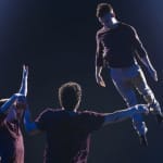 Three aerial acrobats soar through the air in a breathtaking stunt, displaying strength, balance, and synchronisation against a dramatic dark backdrop.