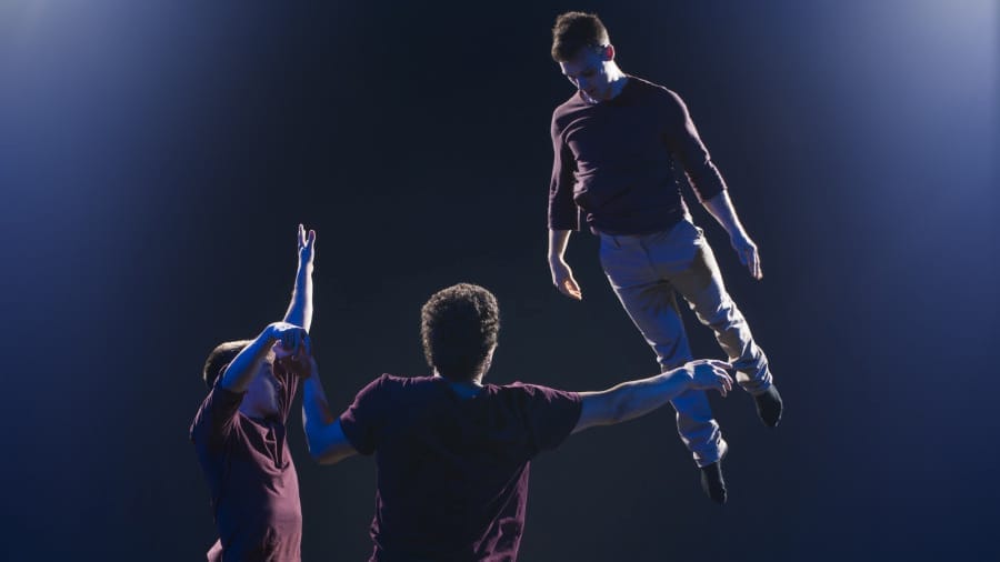 Three aerial acrobats soar through the air in a breathtaking stunt, displaying strength, balance, and synchronisation against a dramatic dark backdrop.