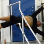 A person performs an acrobatic move on horizontal bars, with both arms extended and legs raised, against a blue door backdrop.