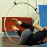 An acrobat performs a dynamic aerial trick on a hoop suspended from the ceiling, while another person observes in a rehearsal space.