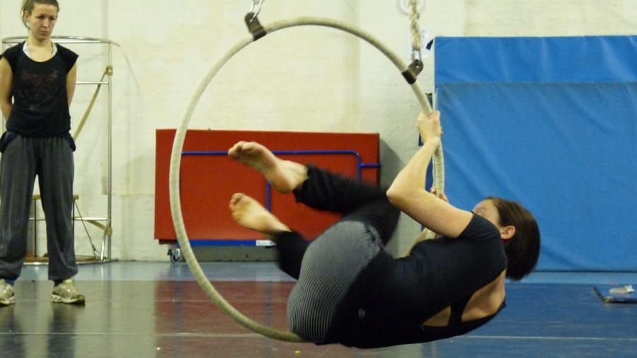 An acrobat performs a dynamic aerial trick on a hoop suspended from the ceiling, while another person observes in a rehearsal space.