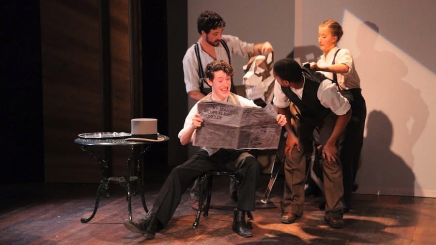 A group of four performers on stage interacting, with one person seated in a chair reading a newspaper in a theatrical setting.