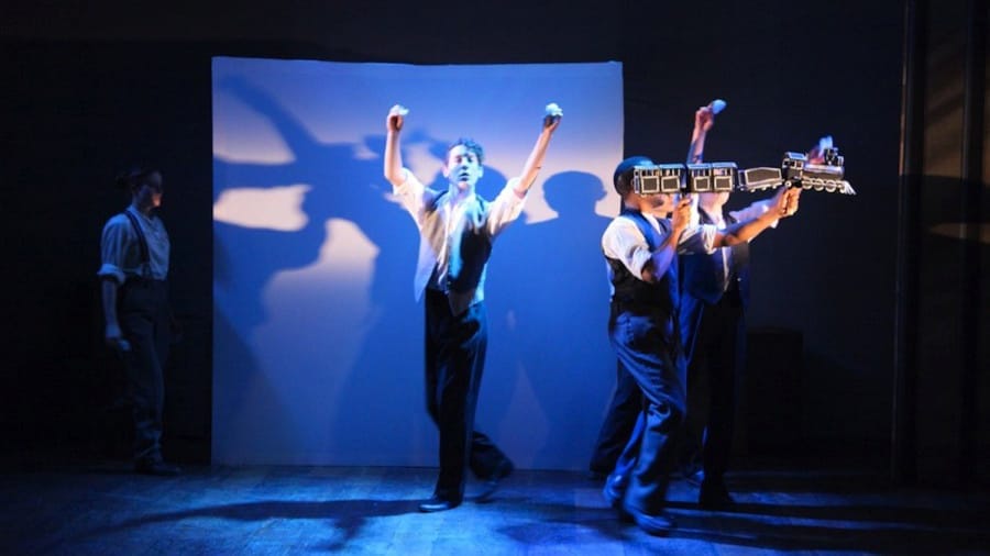 Four performers in vintage attire create dynamic shadows on a blue-lit backdrop, expressing energy through movement and props.