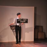 A performer stands on stage against plain white backdrop holding a sign that reads "A FEW YEARS EARLIER," with a basket in hand and wooden boxes in the background.