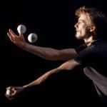 A juggler with brown hair, dressed in black t-shirt, skilfully balances three white balls in motion against a dark background.