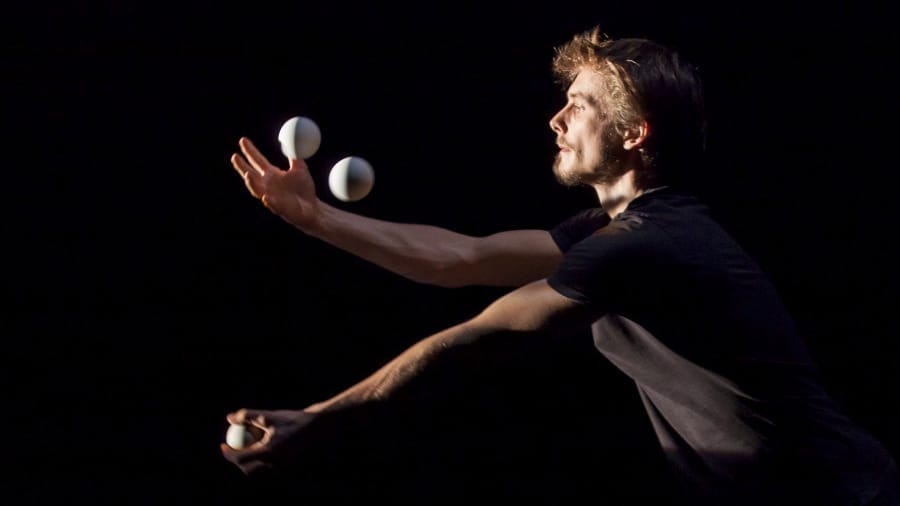 A juggler with brown hair, dressed in black t-shirt, skilfully balances three white balls in motion against a dark background.