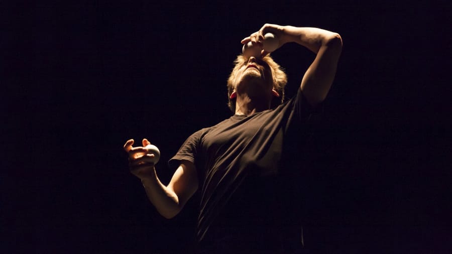 A performer with brown hair, dressed in a black shirt, skilfully juggles three white balls, balancing one on his forehead against a deep black background, showcasing his concentration and skill.