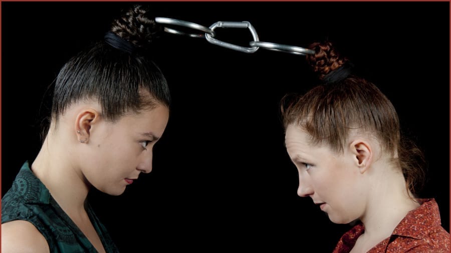 The contrasting hairstyles of two women, connected by a metal chain, create a striking visual image against a dark background.