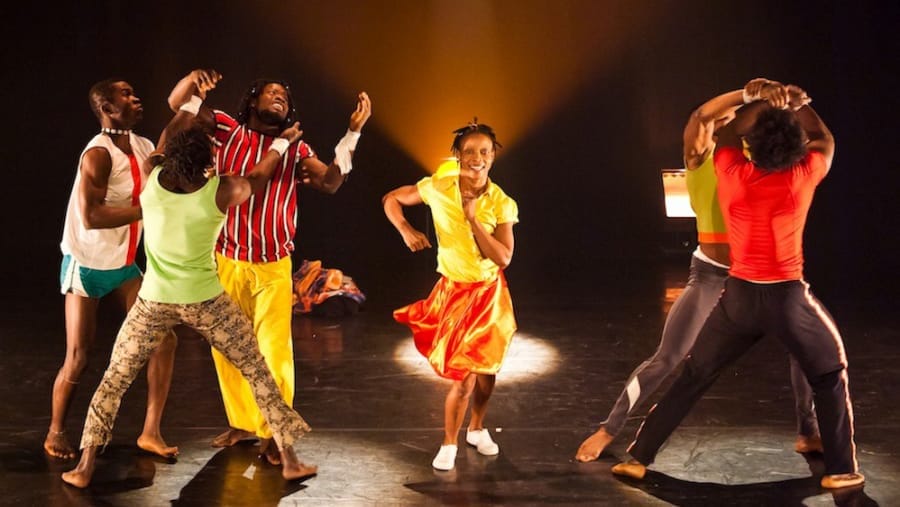 A group of six dancers in colourful outfits perform energetically on stage, under warm stage lighting. One performer in a bright yellow skirt takes centre stage, leading the movement.