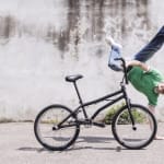 A performer in a green shirt executes a handstand, with one hand on the ground and the other holding their bike, set against a textured wall.