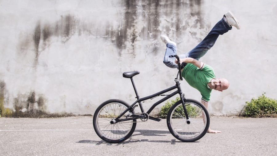 A performer in a green shirt executes a handstand, with one hand on the ground and the other holding their bike, set against a textured wall.
