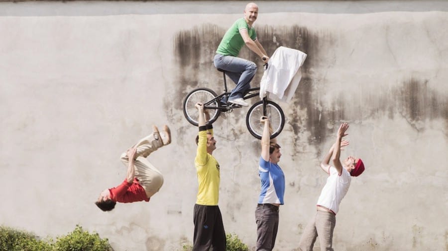A group of five people performs an acrobatic stunt with a bike: one person is mid-air on the bike, two others are holding the bike, one is jumping in mid-air, and another is clapping their hands.