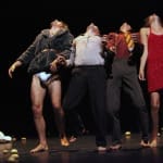 Four performers strike dramatic poses with their heads thrown back and hands reaching forward on stage, surrounded by scattered potatoes against a dark background.
