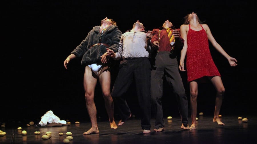 Four performers strike dramatic poses with their heads thrown back and hands reaching forward on stage, surrounded by scattered potatoes against a dark background.
