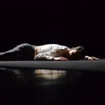 A dancer in black trousers and a white shirt lies flat on a dark stage, illuminated by soft light, creating a dramatic contrast with the shadows around.