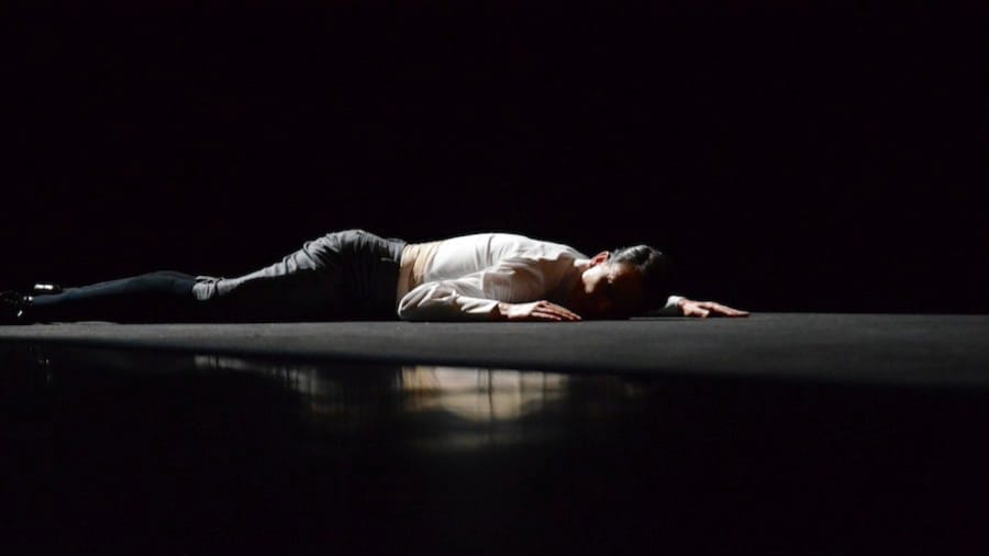 A dancer in black trousers and a white shirt lies flat on a dark stage, illuminated by soft light, creating a dramatic contrast with the shadows around.