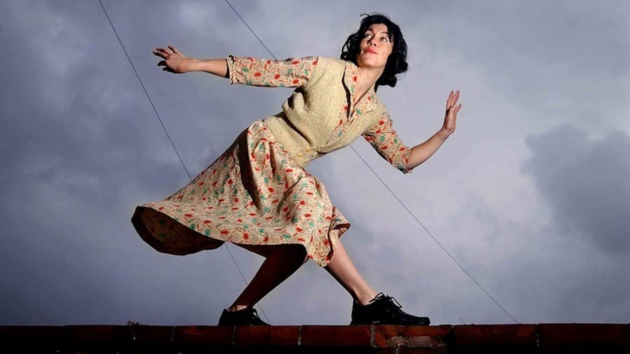 A woman in a vintage floral dress striking a dynamic pose on a rooftop, appearing as if caught mid-motion, with a moody sky behind her.