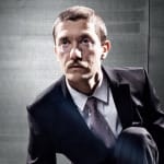A man in a dark suit and tie crouching, looking upwards with a serious expression, against a grey industrial background.