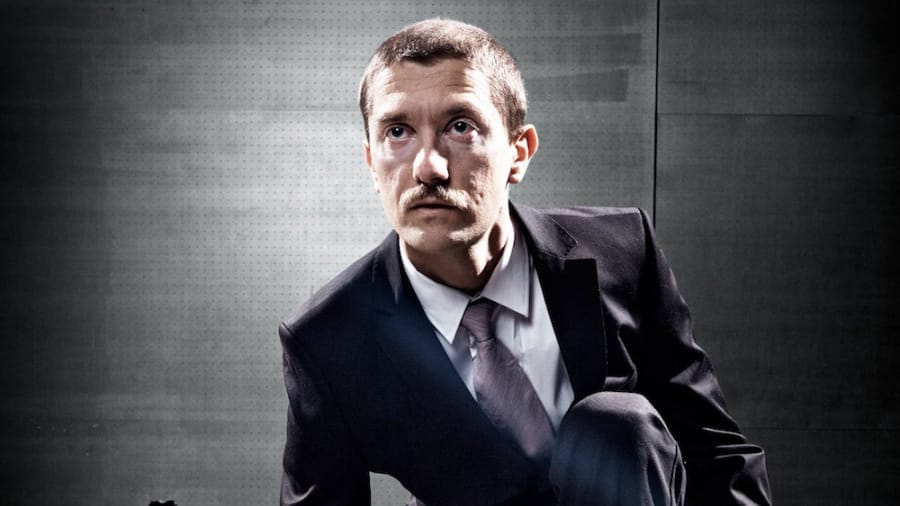 A man in a dark suit and tie crouching, looking upwards with a serious expression, against a grey industrial background.