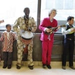 A diverse group of musicians playing various instruments, including a ukulele, drums, and a banjo, in a bright, airy space.