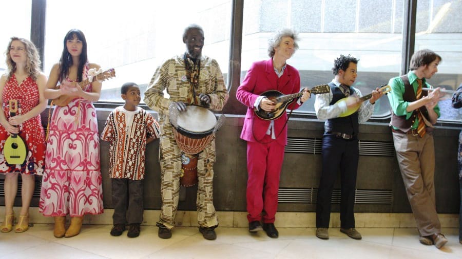 A diverse group of musicians playing various instruments, including a ukulele, drums, and a banjo, in a bright, airy space.