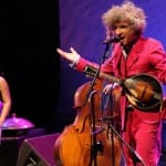 A lively music performance featuring a singing musician in a bright pink suit with a mandolin, accompanied by a violinist in a flower-patterned dress.