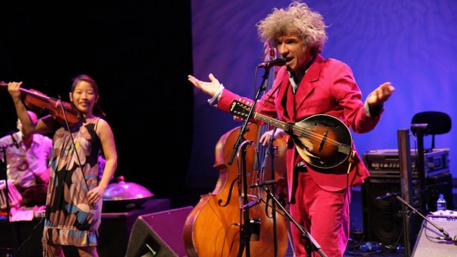 A lively music performance featuring a singing musician in a bright pink suit with a mandolin, accompanied by a violinist in a flower-patterned dress.