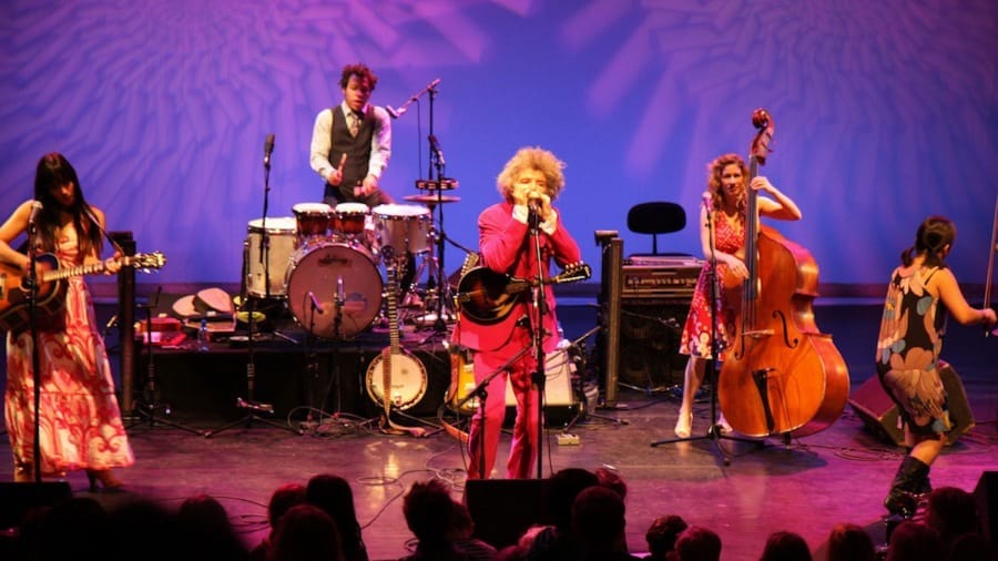 A lively band of five musicians performs on stage with musical instruments, surrounded by an engaged audience in a vibrant, illuminated setting.