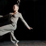 Two acrobats in grey suits move mid-air, suspended by their hair from metal rings on a dark stage.