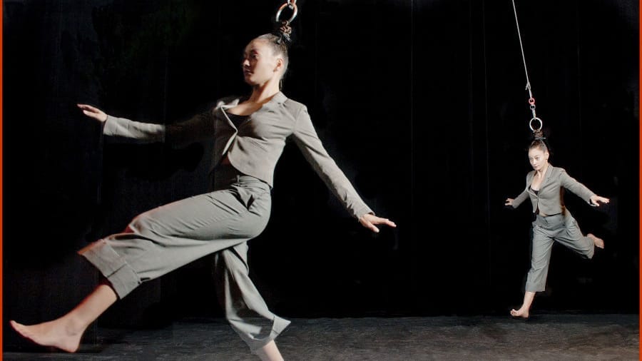 Two acrobats in grey suits move mid-air, suspended by their hair from metal rings on a dark stage.
