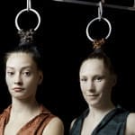 A close-up of two performers standing side by side, their hair attached to metal rings, gazing calmly at the camera against a dark backdrop.