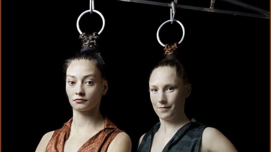 A close-up of two performers standing side by side, their hair attached to metal rings, gazing calmly at the camera against a dark backdrop.