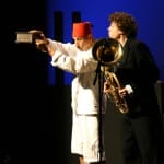 A performer in a white outfit and red fez holds an object while a musician in a black suit plays a tuba on stage.