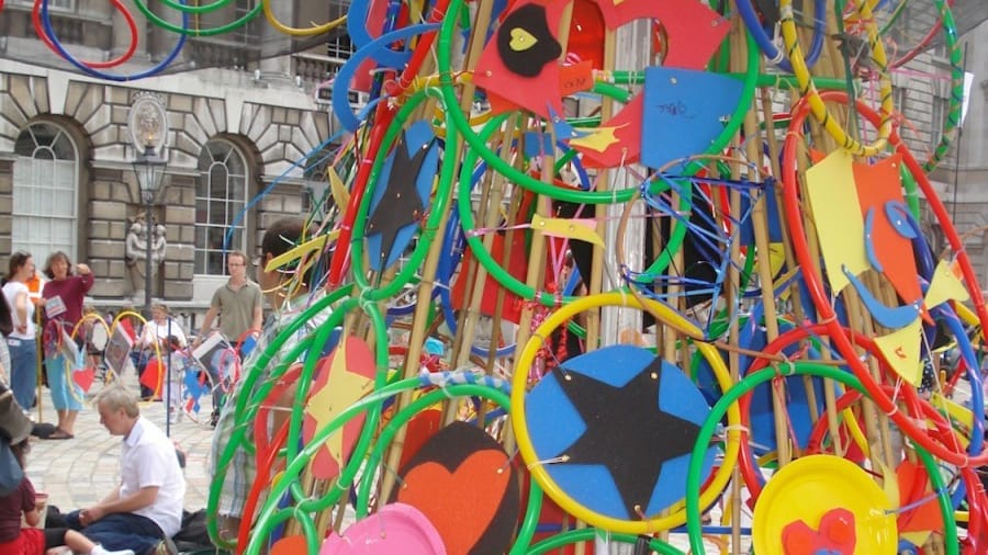 A colourful art installation made of circular hoops and painted shapes, displayed in a courtyard with people sitting on the ground nearby.