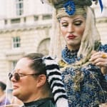 A performer in an extravagant blue and gold baroque-style costume styles a seated participant’s hair in an outdoor setting, with a historic building and a crowd in the background.