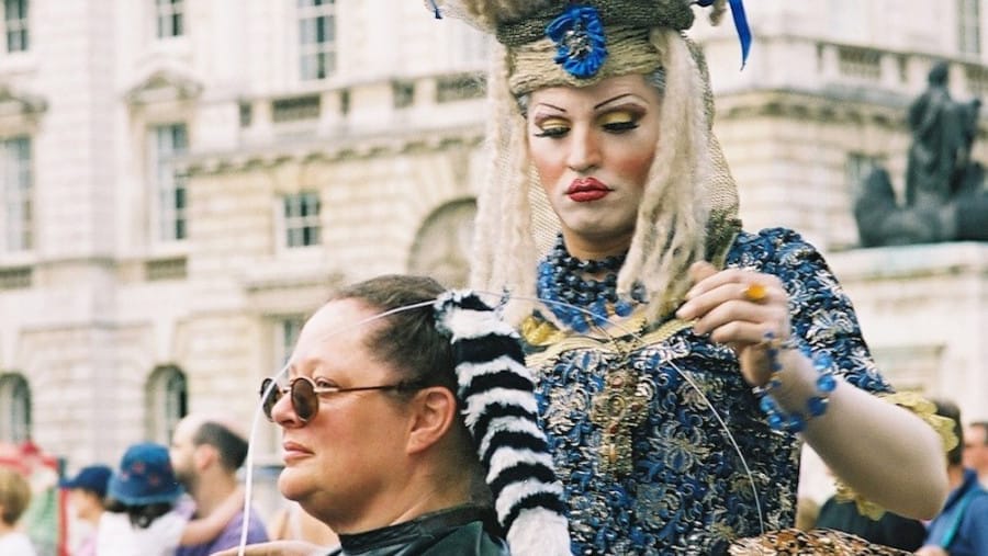 A performer in an extravagant blue and gold baroque-style costume styles a seated participant’s hair in an outdoor setting, with a historic building and a crowd in the background.