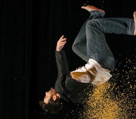 A man in a black jacket and jeans leaps backward, creating a cloud of golden dust against a dark background.