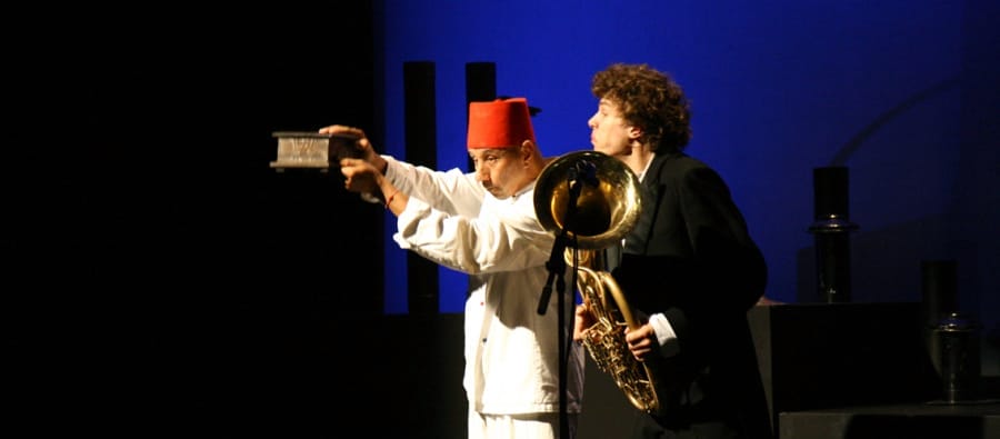 A performer in a white outfit and red fez holds a small box while a musician in a black suit plays a tuba on stage.