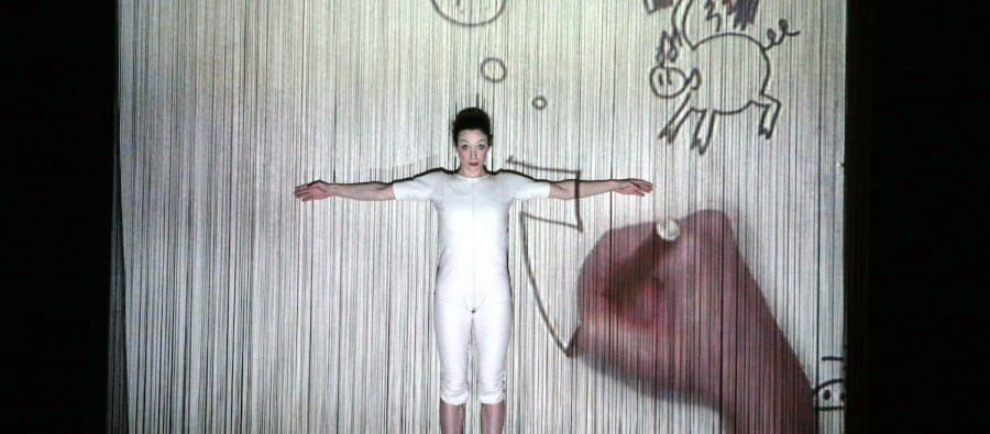 A performer dressed in a white bodysuit stands against a curtain-like screen, arms outstretched, as hand-drawn animated illustrations, including a flying pig and a question mark, are projected onto the backdrop. The image creates a playful and imaginative visual effect, blending live performance with animation. The Great Indoors Manchester